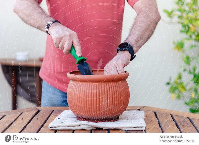 Mann bereitet eine Topfpflanze auf einem sonnigen Hinterhoftisch vor Garten Verguss Pflanze Terrakotta hölzern Tisch im Freien Kelle Gartenarbeit Hobby Tag