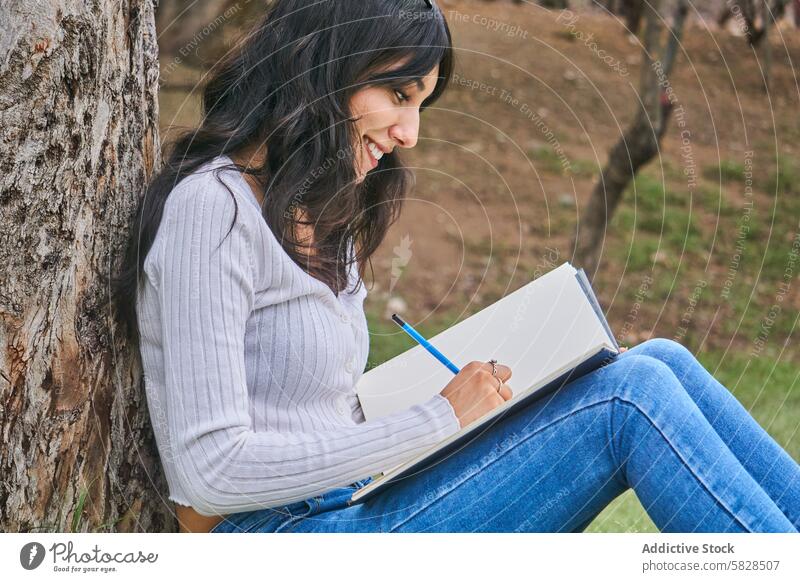 Gelassene Frau beim Skizzieren im Freien unter einem Baum skizzierend Park Gelassenheit sitzend Freizeit Zeichnung entspannend Bleistift Notebook kreativ