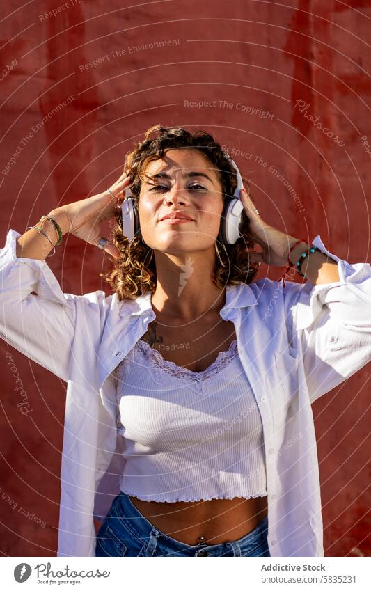 Junge Frau genießt Musik mit Kopfhörern vor einer roten Wand rote Wand freudig krause Haare Freizeitkleidung weißer Kopfhörer Textur junger Erwachsener hören