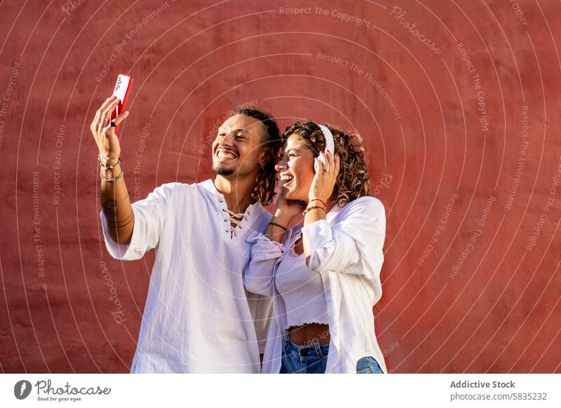 Junges Paar macht Selfie mit einem Smartphone vor einer roten Wand Hintergrund jung heiter Lächeln freudige lässig Outfit weiß spielerisch Glück Sommer sonnig
