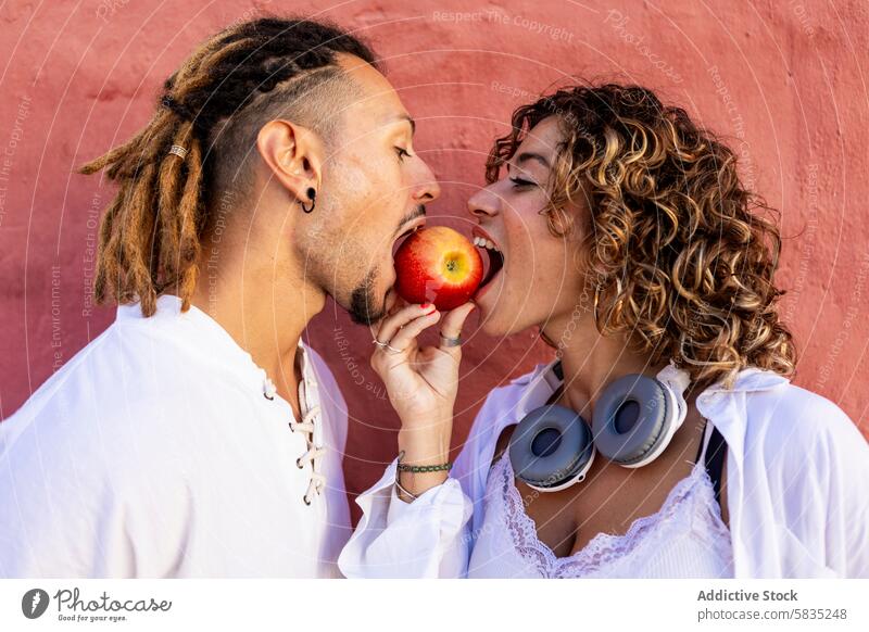 Junges Paar teilt sich einen Apfel vor einer bunten Wand jung spielerisch Biss Mann Frau Freude rote Wand farbenfroh lässig Outfit weißes Hemd Rastalocken