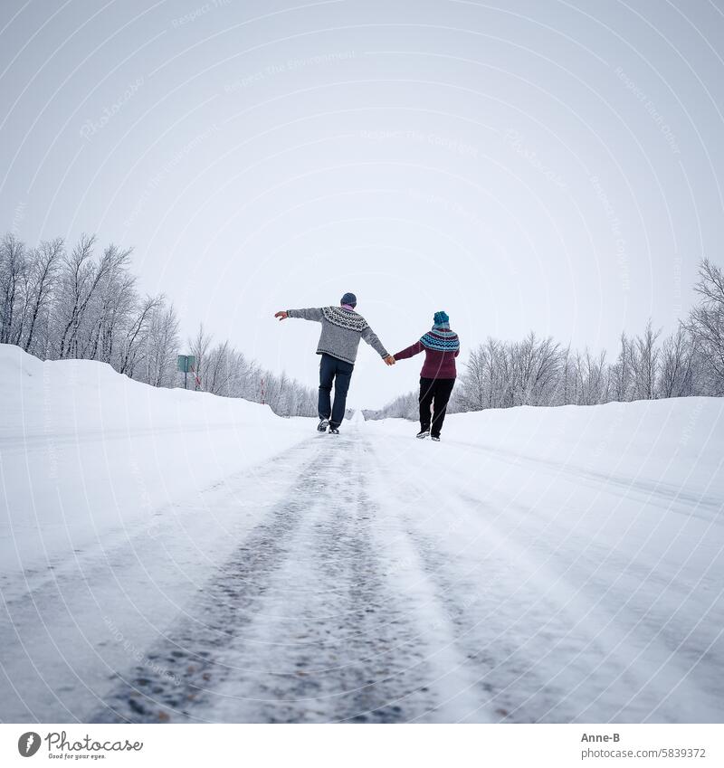 ein fröhliches Paar schlendert Hand in Hand  auf einer Straße in den Nordkalotten im Winter mit schönen dicken selbstgestrickten Pullovern Strickpullover