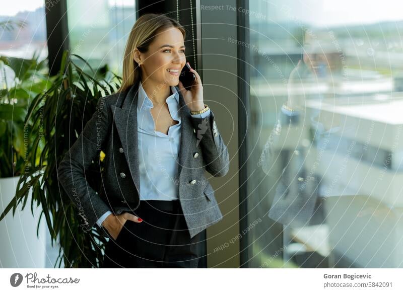 Lächelnde Geschäftsfrau, die bei Tageslicht am Bürofenster mit einem Mobiltelefon spricht Mobile Telefon Gespräch grau Blazer selbstbewusst