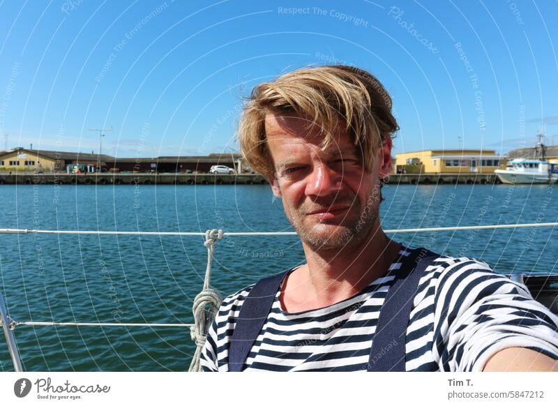 ein Mann auf einem Boot im Hafen Dänemark Matrosenhemd Wasser Außenaufnahme maritim Segeln blond Farbfoto Frühling Segelboot Reeling Ostsee Himmel Bornholm