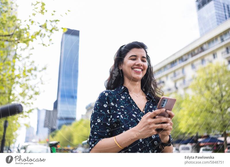 Lächelnde junge Frau benutzt ihr Smartphone in der Stadt Inder echte Menschen Großstadt Spaß Sommer genießend Straße reisen im Freien urban Erwachsener