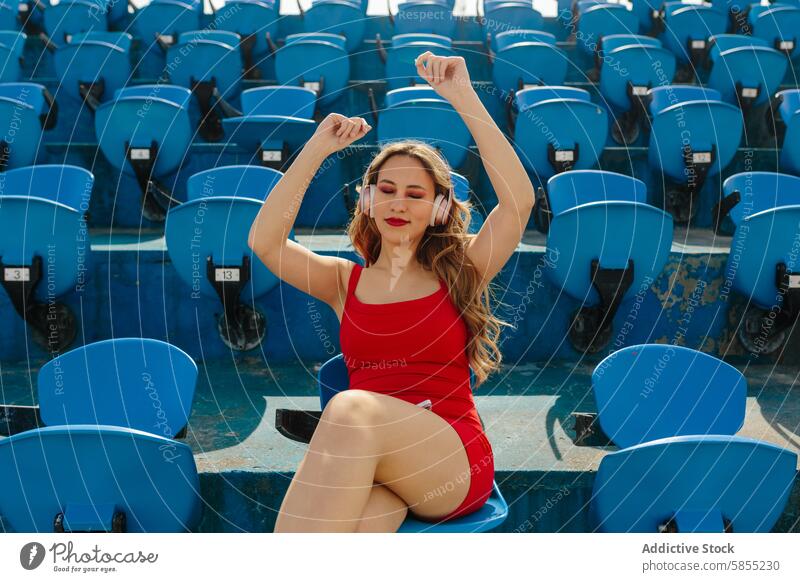 Junge Frau genießt Musik allein auf den Sitzen im Stadion Kopfhörer Feier rot Outfit blau Sitzgelegenheit Entertainment Freizeit Erholung Freude hören im Freien