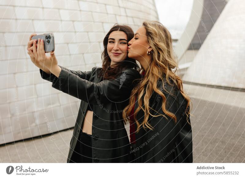 Zwei Frauen machen ein Selfie vor einem modernen Hintergrund Freund Smartphone Kuss Wange Freude offen Moment jung Glück Teilen Genuss urban Mode Stil Lächeln