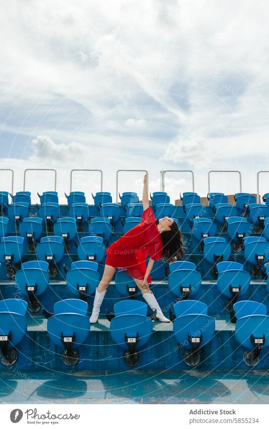 Junge Frau tanzt freudig in einem Stadion Tanzen Sitz rot Kleid blau Himmel Cloud Freude Energie jugendlich Bewegung im Freien Feier Mode Stil Glück pulsierend