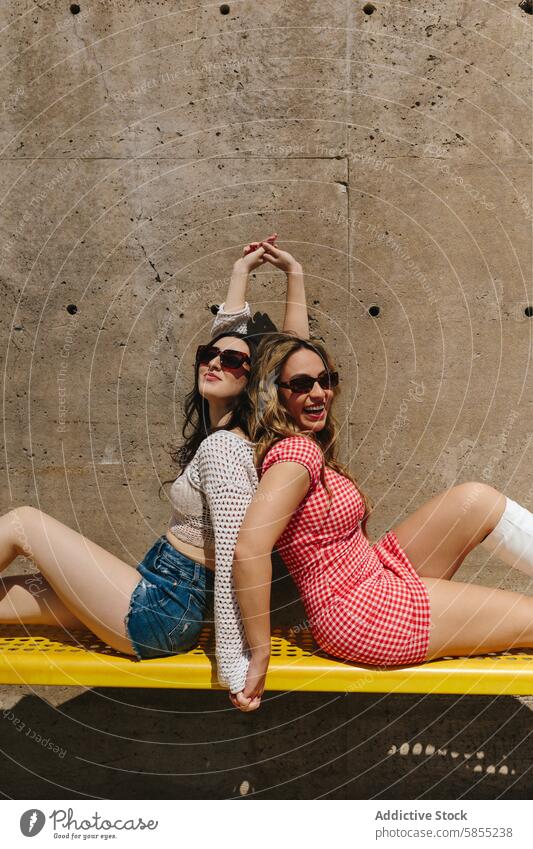 Verspielte Freunde, die einen sonnigen Tag im Park genießen Frau Sommer Spaß Freude heiter Sonnenbrille Kleid Jeansstoff jung Bank gelb im Freien lässig Mode