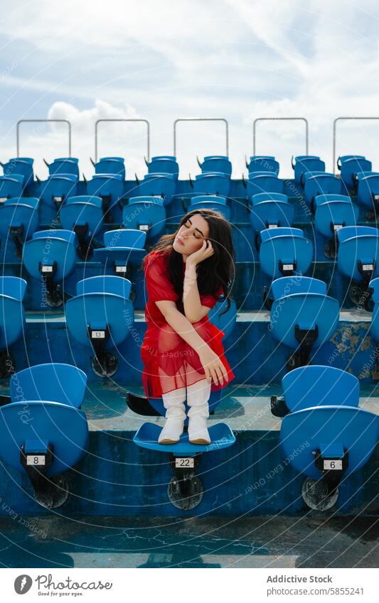 Junge Frau in rotem Kleid sitzt allein auf den Sitzen im Stadion rotes Kleid blauer Sitz nachdenklich Tag im Freien Einsamkeit jung Mode Stil Stiefel