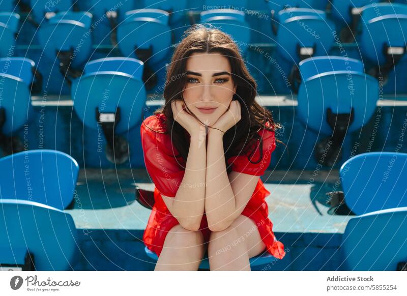 Junge Frau in Rot sitzt allein auf den Sitzen im Stadion rot Kleid Starrer Blick pulsierend Fotokamera jung Mode im Freien tagsüber heiter nachdenklich