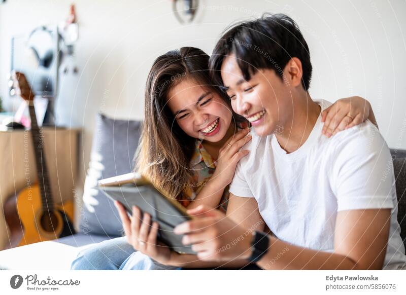 Glückliches junges Paar, das gemeinsam auf dem Sofa zu Hause ein digitales Tablet benutzt asiatisch echte Menschen Spaß genießend junger Erwachsener authentisch