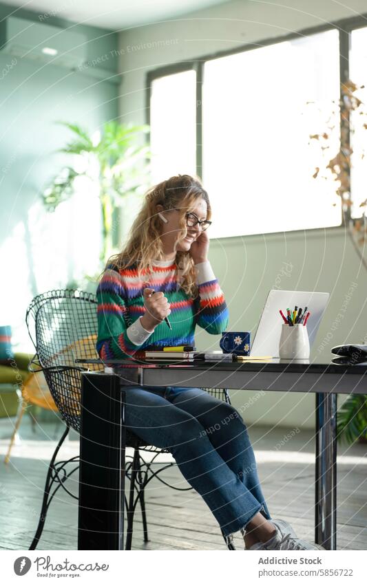 Junge Frau lächelt während einer Fernarbeitssitzung zu Hause Heimarbeitsplatz Laptop Lächeln jung Brille bunter Pullover Schreibtisch Gespräch
