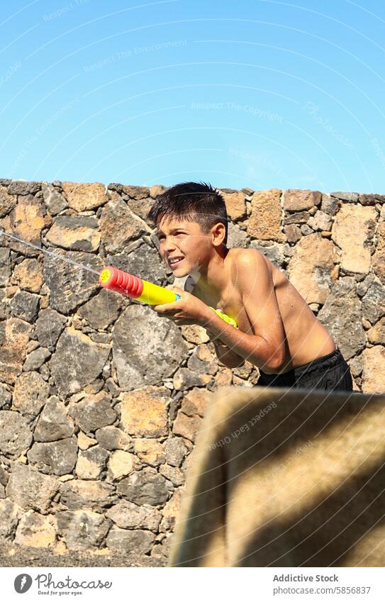 Junge spielt mit Wasserpistole an einer Steinmauer spielen Lächeln Wegsehen Steinwand im Freien sonnig Sommer Spaß Freizeit Aktivität Kindheit Spritzer nass