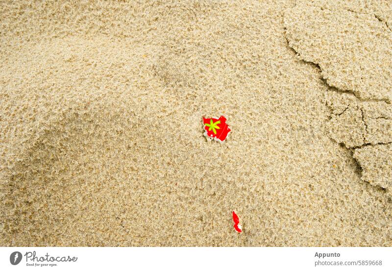 Strandschnipsel am Strand der Träume -  Auf einem Sandstrand liegen kleine rote Papierschnipsel. Auf einem davon sieht man eine stilisierte grüne Pflanze, wobei es sich um eine Palme oder eine Hanfpflanze handeln könnte; Symbolbild