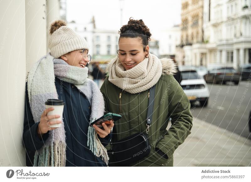 Zwei Frauen genießen ein Gespräch mit Smartphones in London Straße Kaffee Lächeln Talkrunde urban Konnektivität jung Winter Schal Jacke Großstadt lässig