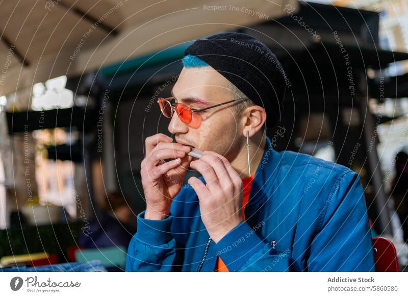 Junger Mann isst Hamburger in Straßencafé mit stilvollem Aussehen jung Essen Café stylisch blau Jacke blaugrün Behaarung schwarz Beanie orange Brille im Freien