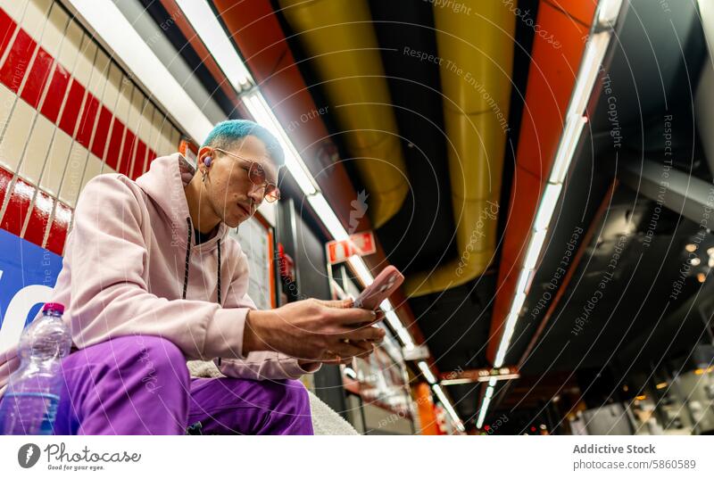 Junger Mann mit blauen Haaren benutzt sein Smartphone in einer U-Bahn-Station jung Sitzen Behaarung purpur Outfit farbenfroh Podest urban Arbeitsweg