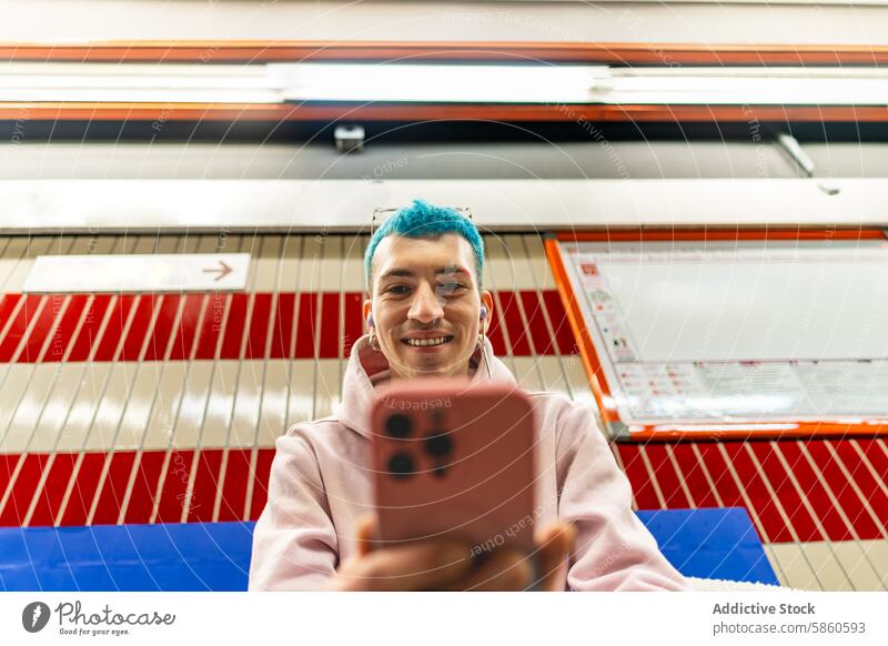 Junger Mann mit blauen Haaren benutzt Smartphone in einer U-Bahn-Station jung blaue Haare Selfie heiter farbenfroh Muster Hintergrund urban Verkehr Pendler