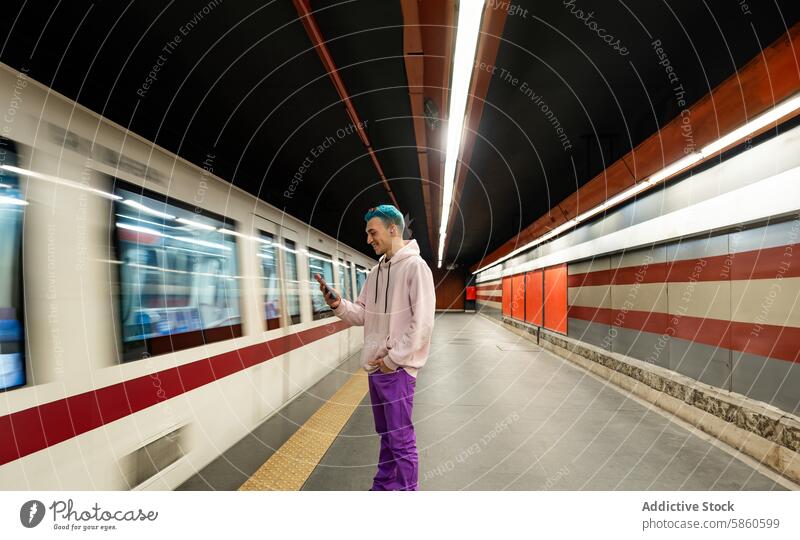 Junger Mann mit blauen Haaren benutzt Smartphone in einer U-Bahn-Station jung blaue Haare Warten Zug Jeansstoff Jacke Im-Ohr-Gerät Kopfhörer Mobile Telefon
