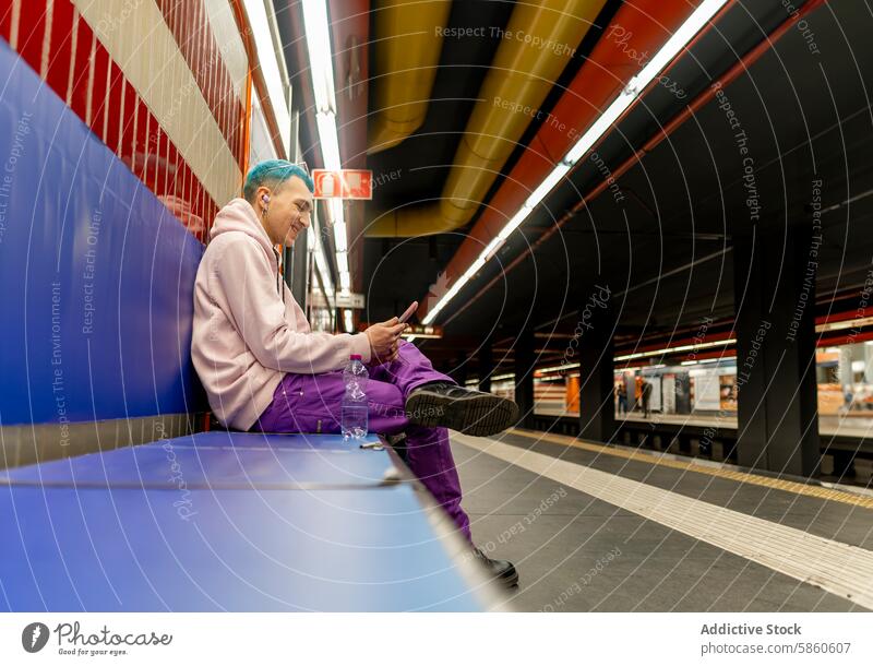 Junger Mann mit blauen Haaren benutzt sein Smartphone in einer U-Bahn-Station jung Sitzen Behaarung purpur Outfit farbenfroh Podest urban Arbeitsweg