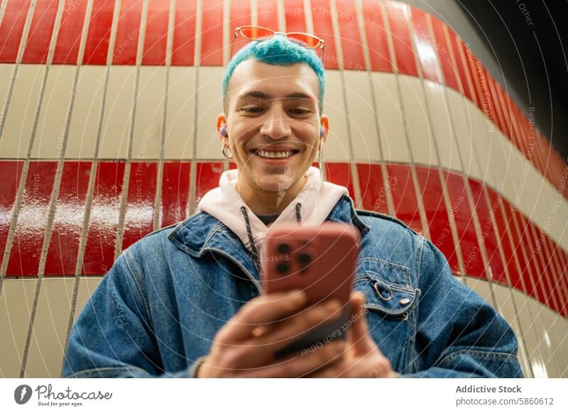 Junger Mann mit blauen Haaren benutzt Smartphone in der U-Bahn blaue Haare Lächeln jung rote Fliese Wand freudig Jeansjacke Brille Glück urban Lifestyle lässig