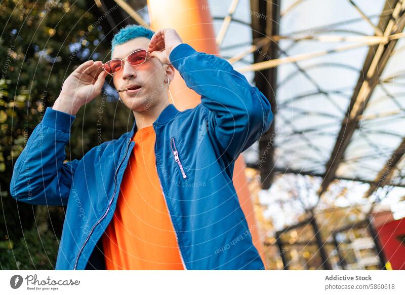 Stilvoller junger Mann mit blauem Haar, der seine Sonnenbrille im Freien verstellt Mode blaue Haare urban modern orangefarbenes Hemd blaue Jacke modisch Brille