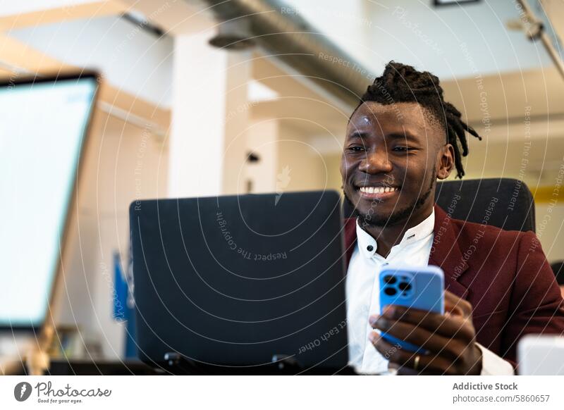 Junge Berufstätige lächelnd mit Smartphone und Laptop professionell Mann jung schwarz Lächeln Büro modern stylisch Blazer rot Multitasking digital Konnektivität