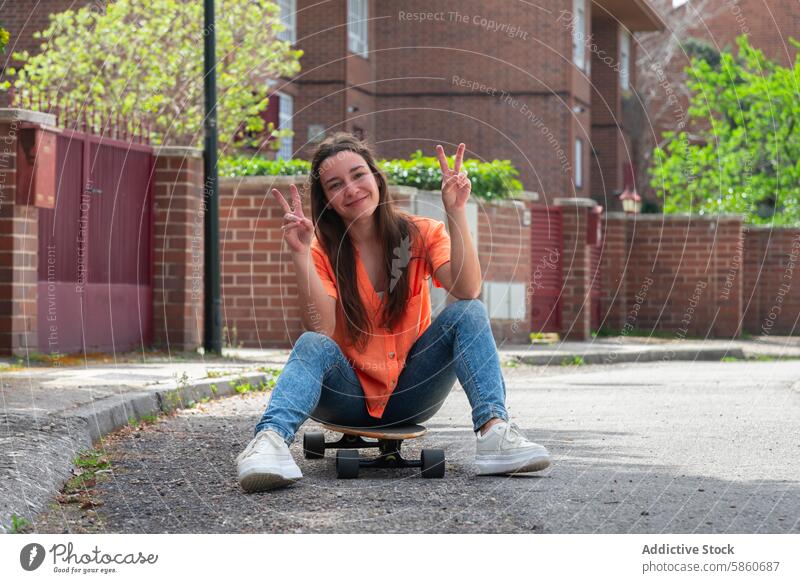 Lächelnde junge Frau, die auf einem Skateboard sitzt und Friedenszeichen macht orangefarbenes Hemd Jeanshose Sitzen Fröhlichkeit Vorstadt Bürgersteig im Freien