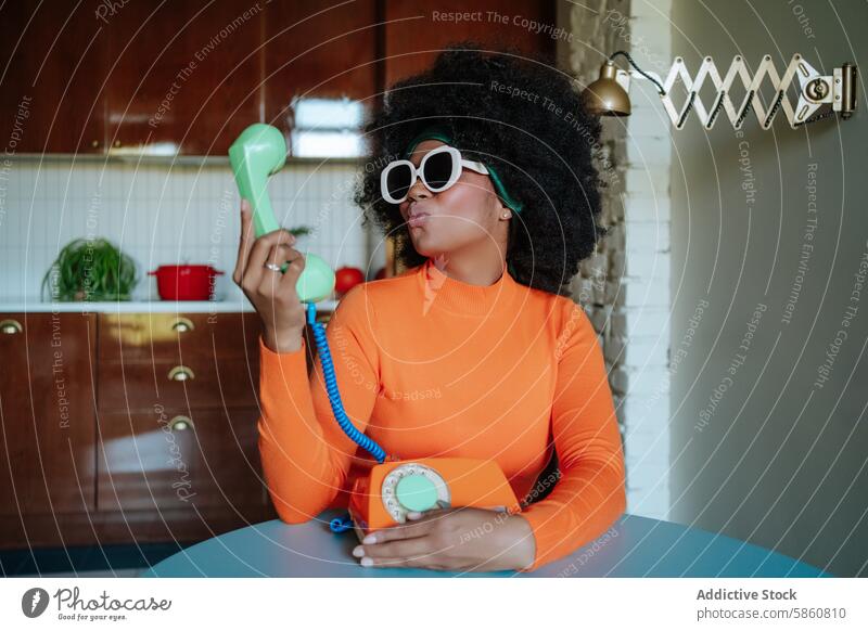 Frau in Retro-Küche küsst ein altes Telefon retro altehrwürdig schwarz 1950s spielerisch Kuss Afro-Look Frisur Sonnenbrille gestylt Hausfrau orange Top Wegsehen