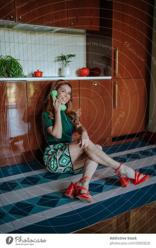 Fröhliche Retro-Hausfrau, die in einer Vintage-Küche am Telefon plaudert altehrwürdig 1950s retro Frau heiter sprechend Sitzen Stock Lachen grün Top gemustert