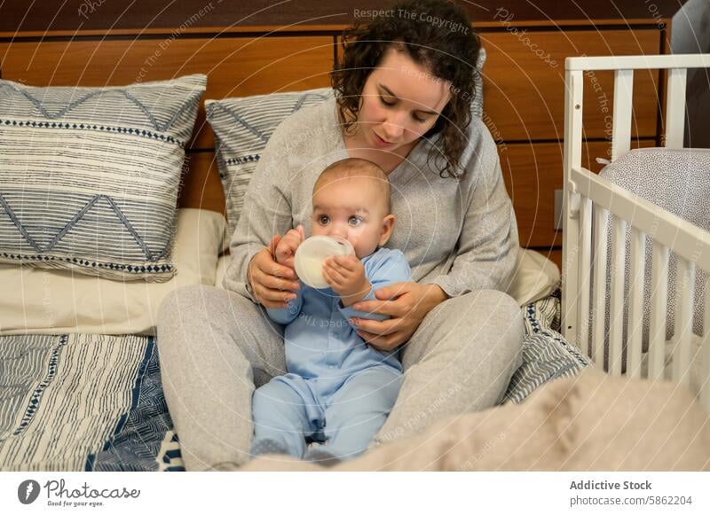 Mutter füttert ihr Baby in einer gemütlichen Schlafzimmerumgebung füttern Flasche Bett Angebot Pflege Pflege von Kaukasier Pyjama bequem blau Kopfkissen