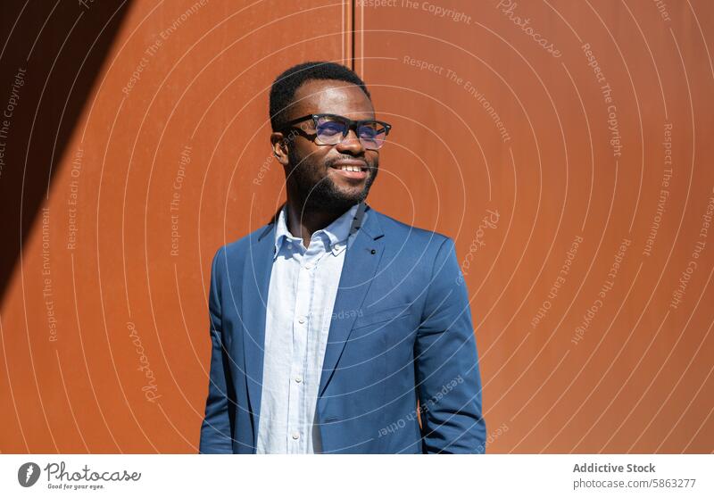 Lächelnder afroamerikanischer Geschäftsmann im blauen Anzug im Freien Afroamerikaner Mann blauer Anzug Brille orangefarbene Wand Seitenansicht professionell