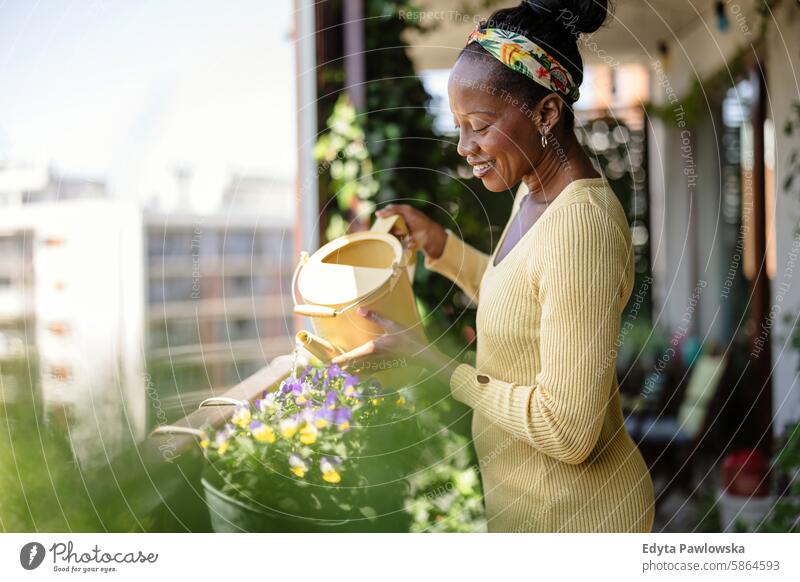 Schöne Frau bewässert Pflanzen auf dem Balkon Menschen Freude schwarz natürlich attraktiv schwarze Frau Fröhlichkeit Glück echte Menschen reif Erwachsener