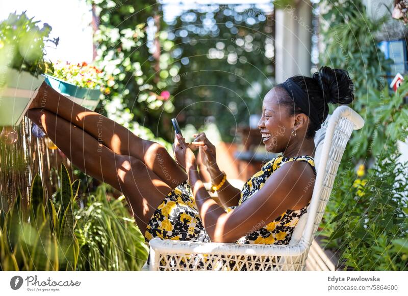 Schöne Frau, die ein Mobiltelefon benutzt, während sie sich auf dem Balkon zu Hause entspannt Menschen Freude schwarz natürlich attraktiv schwarze Frau