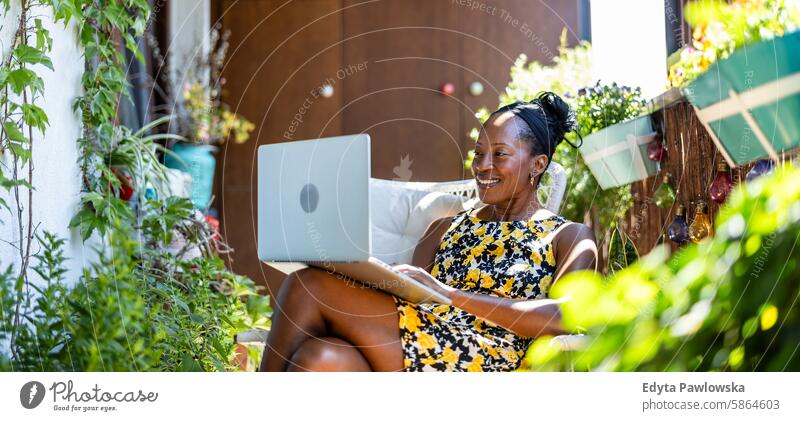Lächelnde Frau mit Laptop auf dem Balkon sitzend Menschen Freude schwarz natürlich attraktiv schwarze Frau Fröhlichkeit Glück echte Menschen reif Erwachsener