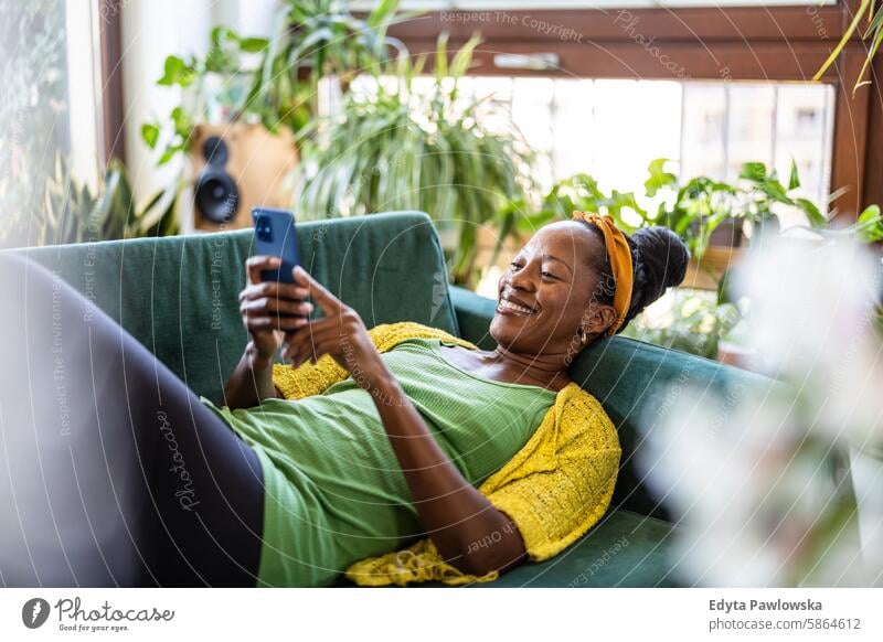 Frau benutzt Smartphone, während sie sich zu Hause auf dem Sofa entspannt Menschen Freude schwarz natürlich attraktiv schwarze Frau Fröhlichkeit Glück