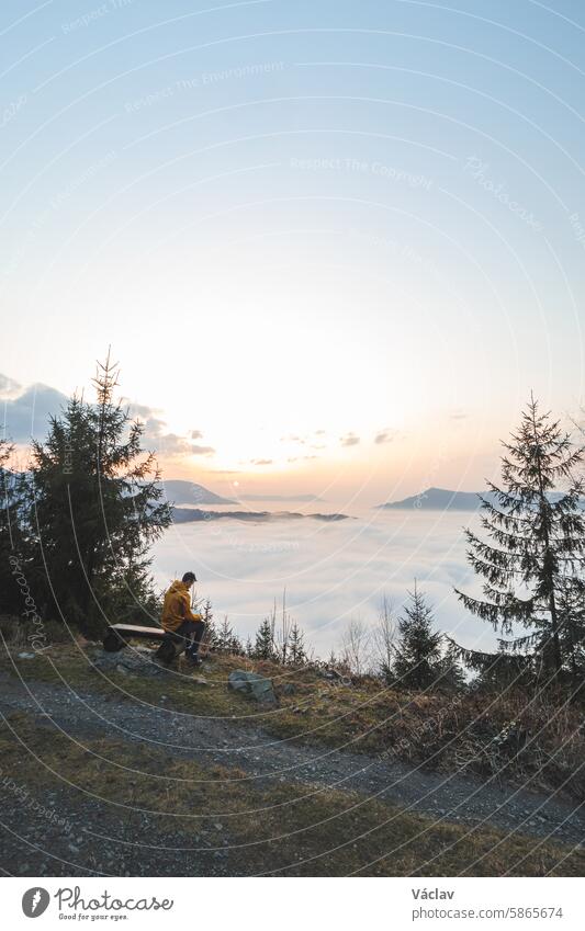 Ein Abenteurer sitzt auf einer Holzbank und beobachtet die Wolken, die unter ihm vorbeiziehen, und die orange-gelben Sonnenuntergänge. Der Mann ist nachdenklich und meditiert. Beskiden-Gebirge