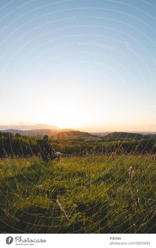 Junger Mann im karierten Hemd sitzt auf einem leichten Reisestuhl und genießt den Blick auf das Tal und die untergehende Sonne am Horizont. Beskiden, Tschechische Republik