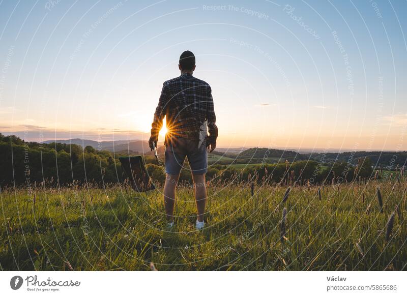 Junger Mann stellt einen Stuhl auf einer Wiese bei Sonnenuntergang auf und macht sich bereit, mit Blick auf das Tal und die untergehende Sonne am Horizont zu entspannen. Beskiden, Tschechische Republik