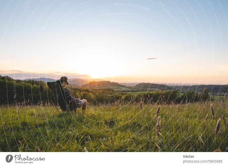 Junger Mann im karierten Hemd sitzt auf einem leichten Reisestuhl und genießt den Blick auf das Tal und die untergehende Sonne am Horizont. Beskiden, Tschechische Republik