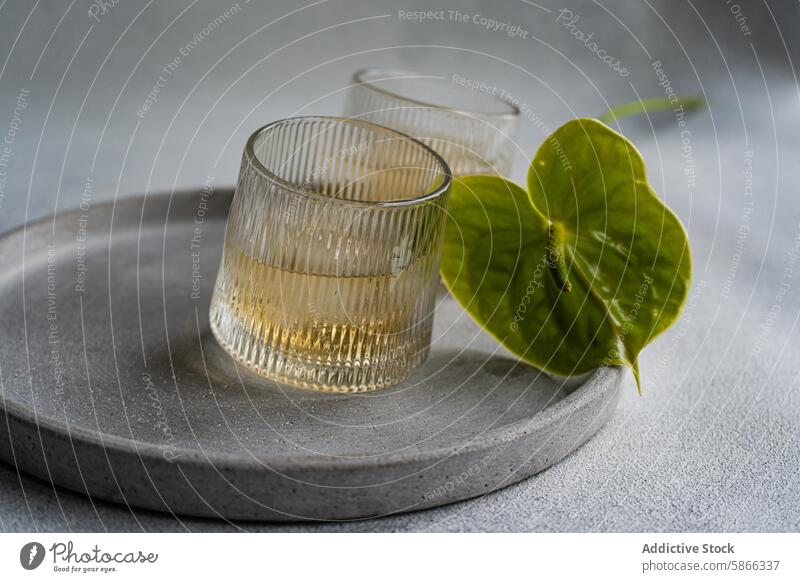 Eleganter Pfirsich-Wodka-Cocktail mit Anthurium auf grauem Teller Vodka Flamingoblumen Glas minimalistisch Tabelleneinstellung Getränk Alkohol trinken Blatt