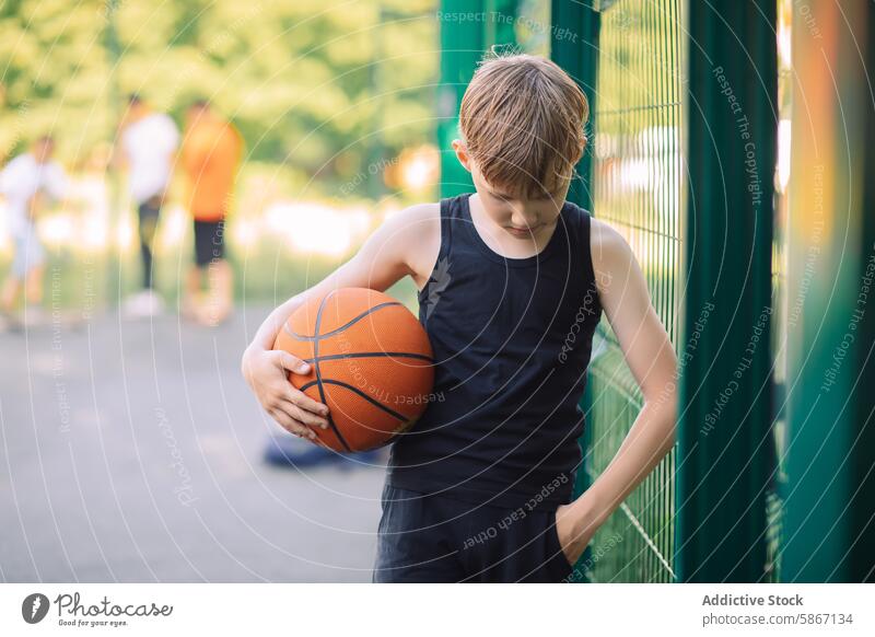 Junger Teenager hält einen Basketball im Freien sportlich Sport Gericht spielen Zaun grün Spiel Freizeit jung Jugend Ball Sommer Halt Stehen lässig Fokus weich