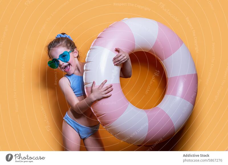 Mädchen mit rosafarbenem Wagen in sommerlichem Studio-Shooting Atelier Sommer Thema Schwimmring Sonnenbrille Badeanzug gelber Hintergrund Freude Spaß pulsierend