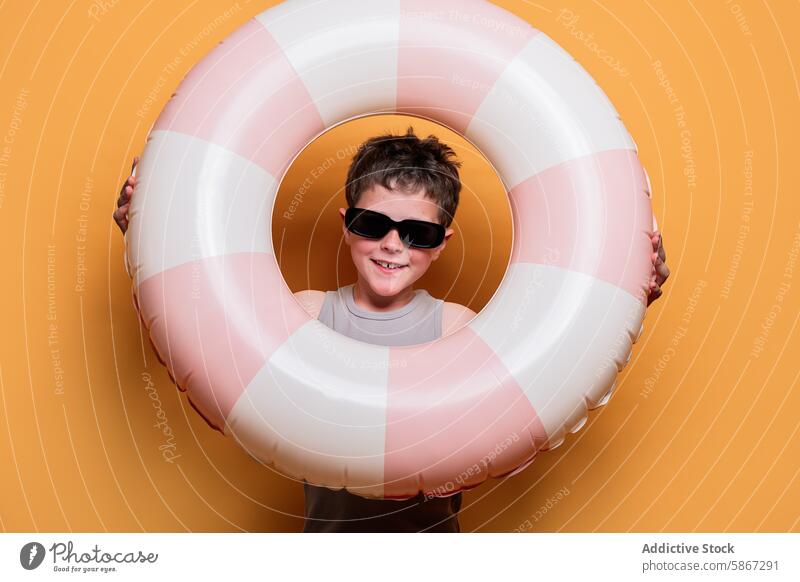 Junge mit Sonnenbrille lächelt durch einen Schwimmring Kind Sommer Atelier Thema Lächeln oranger Hintergrund Fröhlichkeit Freude spielerisch Spaß Freizeit