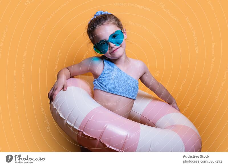 Junges Mädchen mit Sommerrequisiten in Studioumgebung Atelier aufblasbar Schwimmer Sonnenbrille spielerisch orange Hintergrund Kind jugendlich Mode lässig Spaß