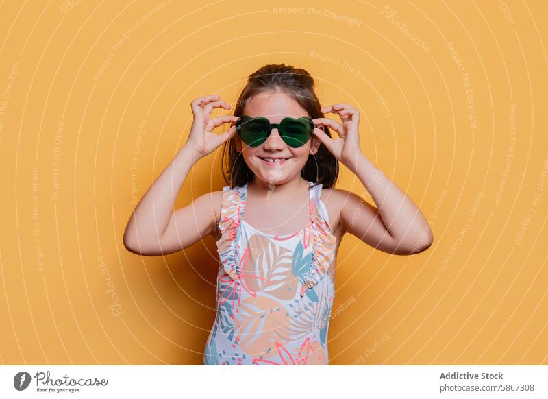 Junges Mädchen in Badeanzug und Sonnenbrille im Studio Kind Atelier Sommer Thema heiter Pose gelber Hintergrund pulsierend tropisch drucken Mode Stil farbenfroh