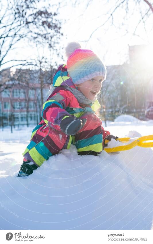 Kind in buntem Schneeanzug spielt im Schnee Spielen farbenfroh Park schaufeln Winter im Freien Aktivität Bäume Gebäude spielerisch jung kalt Umwelt urban