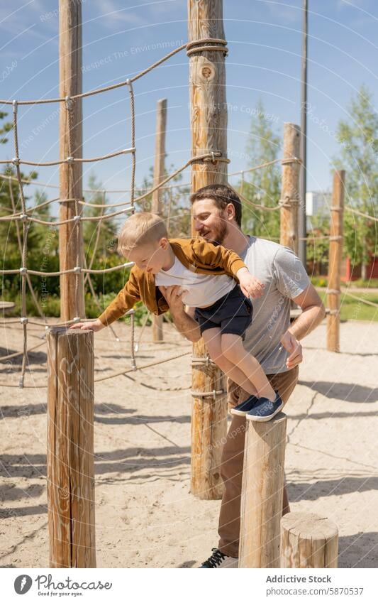 Vater hilft seinem Sohn bei der Navigation durch die Holzspielgeräte im Park Familie Sommer spielen Spielplatz Interaktion abstützen Struktur Gleichgewicht Spaß