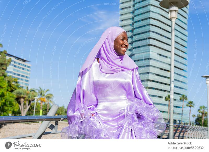 Lächelnde Frau in lila Hidschab genießt den Stadtpark Hijab purpur Park urban Wolkenkratzer strahlend im Freien Mode bescheiden Bekleidung sonnig Tag